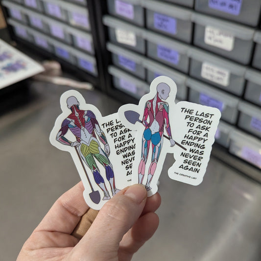 Hand holding three stickers with artistic figures and text in front of a filing cabinet.