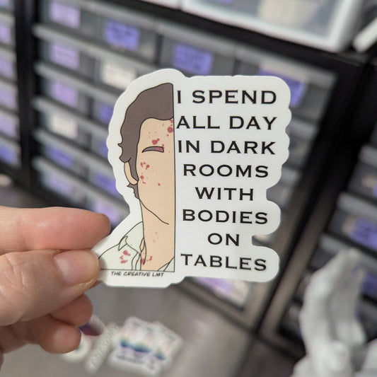 Sticker with text 'I spend all day in dark rooms with bodies on tables' held by a hand in front of a blurred background.